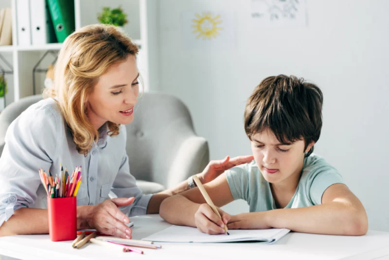 A BCBA sitting beside boy at table, guiding him through a writing assessment with colored pencils.