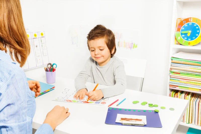 BCBA sits with boy at table during his FBA and watches him draw with colored markers.